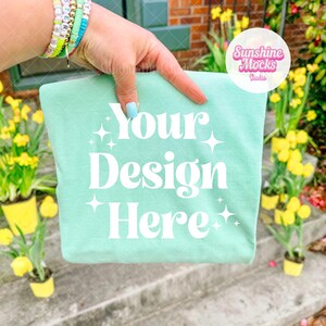 Może przedstawiać: Jasnozielona koszulka z białym napisem "Your Design Here". Koszulka trzymana jest przez dłoń z bransoletkami i jasnoniebieskim pomalowanym paznokciem. Żółte kwiaty w małych doniczkach w tle.