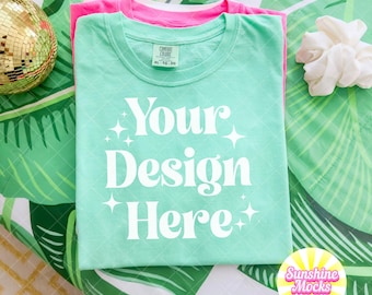 May include: Two folded t-shirts, one pink and one mint green, are stacked on a tropical leaf pattern. The mint green shirt has the text "Your Design Here" in white. A gold disco ball and white scrunchie are also visible.