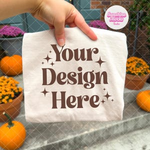 Puede incluir: Una camiseta color crema con el texto "Your Design Here" en marr&oacute;n, con detalles de estrellas. La camiseta se sostiene frente a un fondo borroso con calabazas y flores.