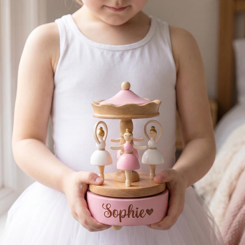 May include: A wooden musical carousel featuring three ballerina figurines in white and pink dresses. The base is painted pink and displays the name Sophie in a cursive font next to a small heart icon. The top canopy is light pink.