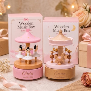 Op de afbeelding: Twee houten muziekdozen met roze en naturel houten afwerkingen. De linkerdoos toont drie ballerina's en de naam Olivia met een datum. De rechterdoos toont een carrouselpaard en de naam Emma met de tekst Happy Birthday.