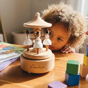 May include: A wooden carousel music box featuring three ballerina figurines in white dresses. The music box has a light wood finish and the name "Amara" is engraved on the base. The carousel is set on a wooden table with colorful blocks.