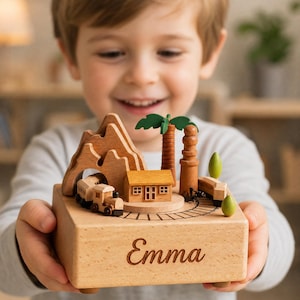 May include: A wooden music box featuring a miniature train set, a small house, a palm tree, and mountain cutouts on top. The base is crafted from light-colored wood and has the name Emma engraved in a cursive script on the front face of the box.