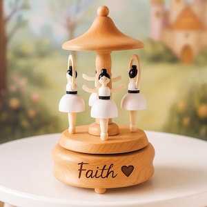 May include: A wooden music box carousel featuring three ballerina figures with black hair and white dresses. The base is made of light-colored wood and has the word Faith engraved in a script font next to a small heart shape symbol on the front.