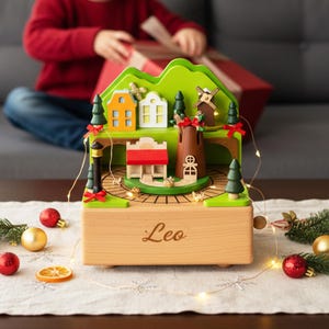 May include: A wooden music box depicting a village scene with a green mountain backdrop, houses, a windmill, and trees. The box is light brown with the name "Leo" engraved on the front. Red and gold baubles and fairy lights surround the box.