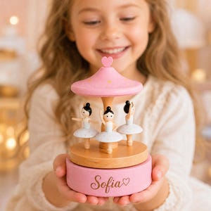 May include: A wooden musical carousel featuring three miniature ballerinas in white tutus. The carousel has a pink roof with a small heart topper and a pink base engraved with the name Sofia and a heart symbol. The structure is made of wood.