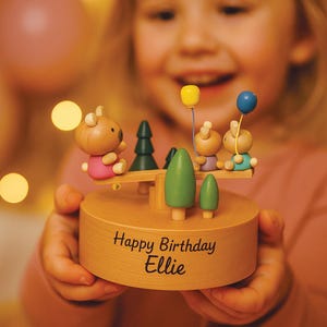 May include: A wooden music box with a seesaw design, featuring bear and bunny figures, trees, and balloons. The box is inscribed with "Happy Birthday Ellie". The figures are painted in pink, blue, and yellow. The music box is held by a person.
