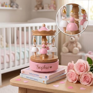 May include: A wooden musical carousel featuring three ballerina figurines in pink and white dresses. The base is pink with the name Sophie and a heart icon printed on it. The carousel sits on two books titled baby barbies and baby board books.