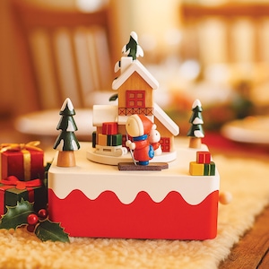 May include: A festive Christmas music box featuring a skier, a house, and miniature trees. The base is red with a white snow-like trim. Presents and a small snowman are also included in this holiday scene.