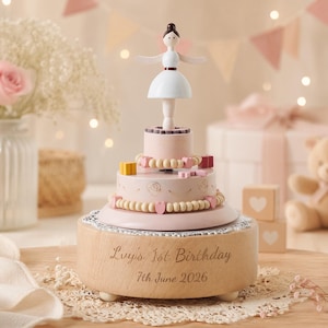 May include: A wooden music box featuring a ballerina figurine on top of a tiered cake design. The base is engraved with the text "Luy's 1st Birthday 7th June 2026". The cake is decorated with small wooden beads, heart shapes, and gift boxes.