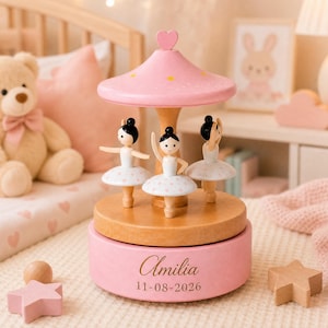 May include: A wooden musical carousel featuring three ballerina figurines in white tutus with pink polka dots. The base is pink with the name Amilia and the date 11-08-2026 engraved in gold. The top is a pink canopy topped with a small heart.