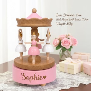 May include: A wooden carousel music box featuring three ballerina figures in pink and white dresses. The pink base is engraved with the name Sophie and a heart. It has a base diameter of 11 cm, a total height of 17.5 cm, and a weight of 385 g.