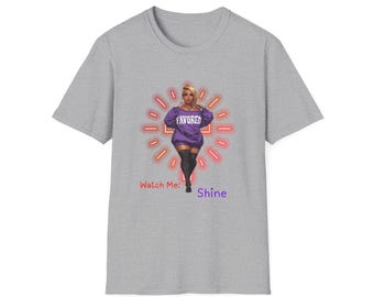 May include: A heather gray t-shirt featuring a graphic of a woman in a purple dress with the word "FAVORED" and the text "Watch Me: Shine". The graphic is surrounded by a neon-style sunburst.