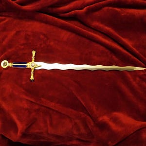 May include: A decorative sword with a wavy blade, gold accents, and a blue handle. The hilt features a cross design with a central jewel. The sword is displayed on a red velvet surface.