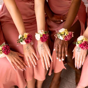 May include: A group of people wearing matching pink dresses, each with a floral wrist corsage. The corsages feature a mix of red, white, and yellow flowers. Each person has a temporary tattoo on their hand with the text "Michelle Patricia".