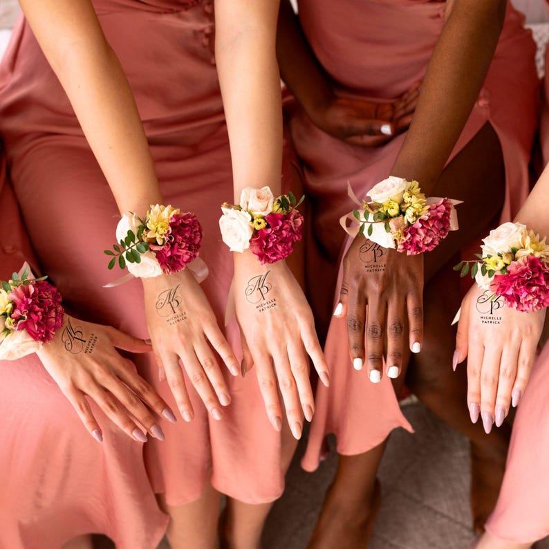 May include: A group of people wearing matching pink dresses, each with a floral wrist corsage. The corsages feature a mix of red, white, and yellow flowers. Each person has a temporary tattoo on their hand with the text "Michelle Patricia".