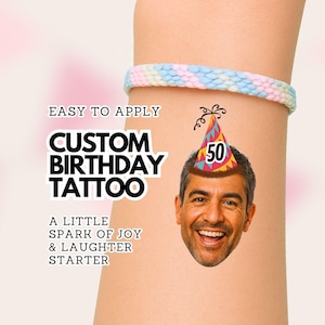 Op de afbeelding: Een tijdelijke tatoeage met een lachend gezicht dat een kleurrijke feesthoed draagt met het getal 50 erop. De tekst luidt EASY TO APPLY CUSTOM BIRTHDAY TATTOO A LITTLE SPARK OF JOY & LAUGHTER STARTER. Een pastelkleurige gevlochten armband is zichtbaar.
