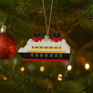 May include: A Christmas tree ornament shaped like a cruise ship, constructed from white, black, red, and yellow toy blocks. The ship has two red and black smokestacks and is suspended by a silver string. The ornament is set against a backdrop of a Christmas tree.
