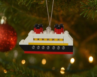 May include: A Christmas tree ornament shaped like a cruise ship, constructed from white, black, red, and yellow toy blocks. The ship has two red and black smokestacks and is suspended by a silver string. The ornament is set against a backdrop of a Christmas tree.