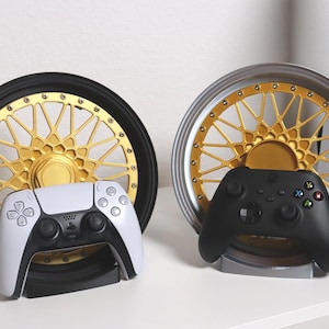 Könnte beinhalten: Zwei Gaming-Controller-Ständer mit einem gold-schwarzen Felgendesign. Einer hält einen weiß-schwarzen PlayStation 5-Controller, der andere einen schwarzen Xbox-Controller. Die Ständer stehen auf einer weißen Oberfläche. Der Raddurchmesser beträgt ca. 30 cm.