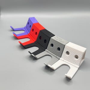 Op de afbeelding: Een rij 3D-geprinte telefoonstandaards in paars, rood, zwart, grijs en wit. Elke standaard heeft een rechthoekige basis met een gebogen steun en een gestructureerd oppervlak. De tekst "Form Kraft 3D" staat op de zwarte en grijze standaards.