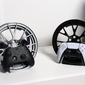 Può includere: Due supporti per controller di gioco. Un supporto argentato tiene un controller Xbox nero. L'altro supporto nero tiene un controller PlayStation bianco e nero. Entrambi i supporti hanno la forma di ruote di auto.