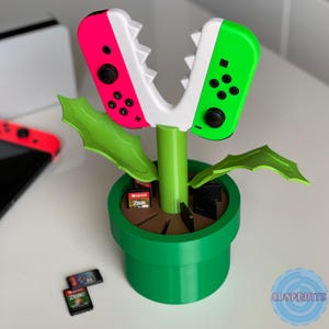 Può includere: Un supporto per controller Switch verde, bianco e rosa a forma di pianta. Il supporto ha un vaso verde, uno stelo verde e foglie verdi. Le impugnature del controller sono rosa e verdi. Diverse cartucce di gioco sono nel vaso.