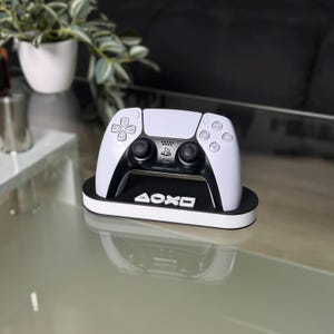 Może przedstawiać: Biało-czarny kontroler do gier wideo spoczywający na czarno-białej podstawce. Podstawka ma biały napis "AOXD". Kontroler ma czarne przyciski i czarny środek.