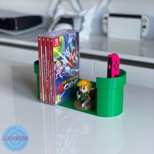 Peut inclure: Un organisateur de jeux vidéo vert imprimé en 3D en forme de tuyau, contenant plusieurs boîtiers de jeux Nintendo Switch, une manette rose et une figurine Link. Les boîtiers de jeux incluent "Super Mario 3D All-Stars" et "Mario Tennis Aces".
