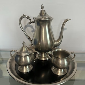 Puede incluir: Un juego de té plateado, que incluye una tetera, un azucarero y una jarra para la crema, presentado en una bandeja redonda. La tetera tiene un asa curva y un pico largo. El juego es de metal y tiene un diseño clásico.