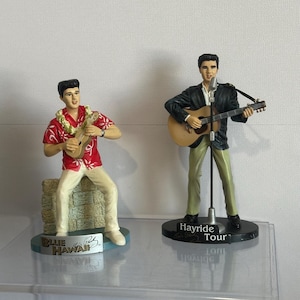 May include: Two figurines of a musician, one playing a ukulele and the other a guitar. The first figure wears a red shirt and white trousers, with a lei. The second figure wears a black jacket and khaki trousers. The base of the first figure says "Blue Hawaii".