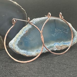 May include: A pair of copper-colored hoop earrings. The earrings have a hammered texture and a simple, circular design. The hoops are displayed against a backdrop of blue and gray stone.
