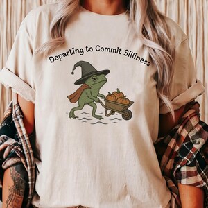 Puede incluir: Camiseta beige con la frase "Departing to Commit Silliness" sobre una rana de dibujos animados con sombrero de bruja y capa, empujando una carretilla de calabazas. La rana es verde, las calabazas son naranjas y el sombrero es negro.