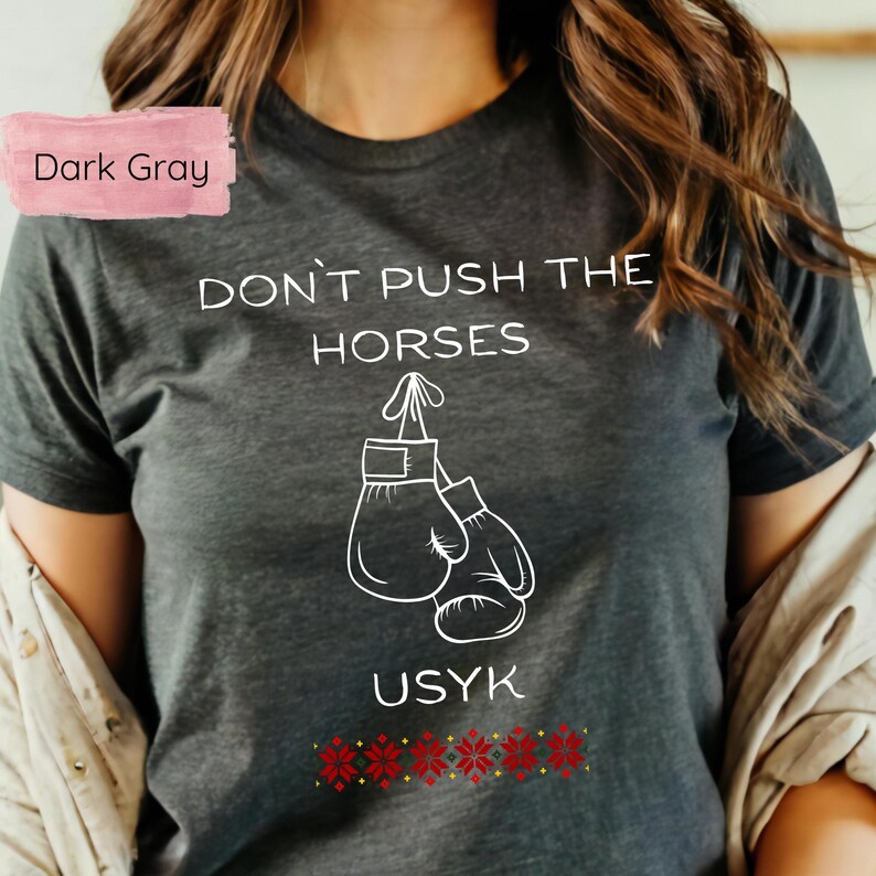 May include: Dark gray t-shirt with the text "DON'T PUSH THE HORSES" and "USYK" in white. The design includes a pair of boxing gloves. A red and gold floral pattern is at the bottom of the shirt.