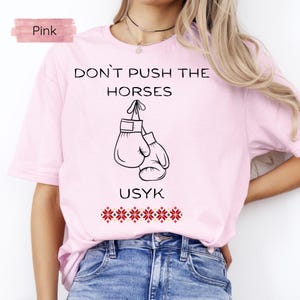 May include: A light pink t-shirt with the text "DON'T PUSH THE HORSES" above a graphic of boxing gloves. Below the gloves is the name "USYK" and a decorative floral pattern.