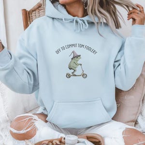 May include: A light blue hooded sweatshirt featuring a graphic of a green frog wearing a pointed hat and riding a scooter. The text above the frog reads, "OFF TO COMMIT TOM FOOLERY." The garment includes a front kangaroo pocket and a drawstring hood.