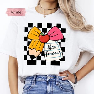 Puede incluir: Camiseta blanca con un dise&ntilde;o tem&aacute;tico para profesores. El dise&ntilde;o incluye un l&aacute;piz amarillo, un lazo rosa, una manzana roja y un bloc de notas con el texto "Mrs. Teacher" sobre un fondo a cuadros blanco y negro.