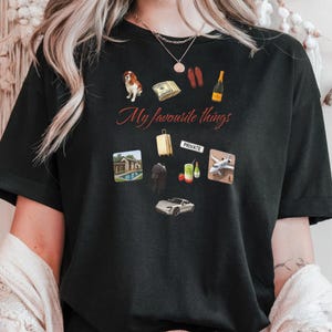 May include: Black t-shirt with the phrase "My favourite things" and various graphic illustrations. The graphics include a dog, money, sandals, champagne, a house with a pool, a suitcase, a private sign, a person, a cocktail, a plane, and a car.