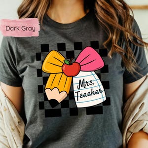 Puede incluir: Camiseta gris oscuro con un dise&ntilde;o gr&aacute;fico colorido. El dise&ntilde;o incluye un l&aacute;piz amarillo, un lazo rosa y una manzana roja. Las palabras "Mrs. Teacher" est&aacute;n escritas en un bloc de notas. El fondo es un patr&oacute;n de cuadros blancos y negros.