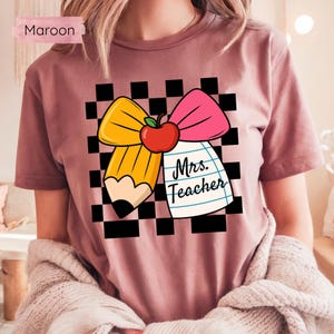 Puede incluir: Camiseta granate con un dise&ntilde;o gr&aacute;fico que presenta un l&aacute;piz amarillo, un lazo rosa y una manzana roja. El dise&ntilde;o incluye el texto "Mrs. Teacher" en un bloc de notas sobre un fondo a cuadros blanco y negro.