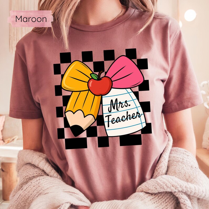 Puede incluir: Camiseta granate con un dise&ntilde;o gr&aacute;fico que presenta un l&aacute;piz amarillo, un lazo rosa y una manzana roja. El dise&ntilde;o incluye el texto "Mrs. Teacher" en un bloc de notas sobre un fondo a cuadros blanco y negro.