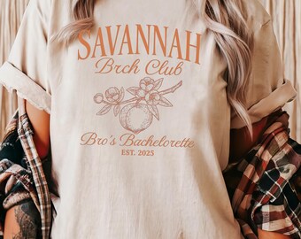 Può includere: Una maglietta color naturale con la scritta "SAVANNAH Brch Club" in arancione. Sotto, un'illustrazione di un ramo di pesca e le parole "Bro's Bachelorette EST. 2025" in arancione. La maglietta ha le maniche arrotolate.