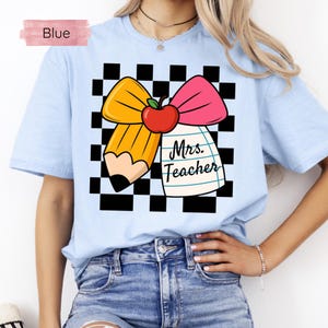 Puede incluir: Camiseta azul claro con un gr&aacute;fico que presenta un l&aacute;piz amarillo, un lazo rosa y una manzana roja sobre un fondo a cuadros blanco y negro. El texto "Mrs. Teacher" est&aacute; impreso en un bloc de notas. La palabra "Blue" est&aacute; en un rect&aacute;ngulo rosa.
