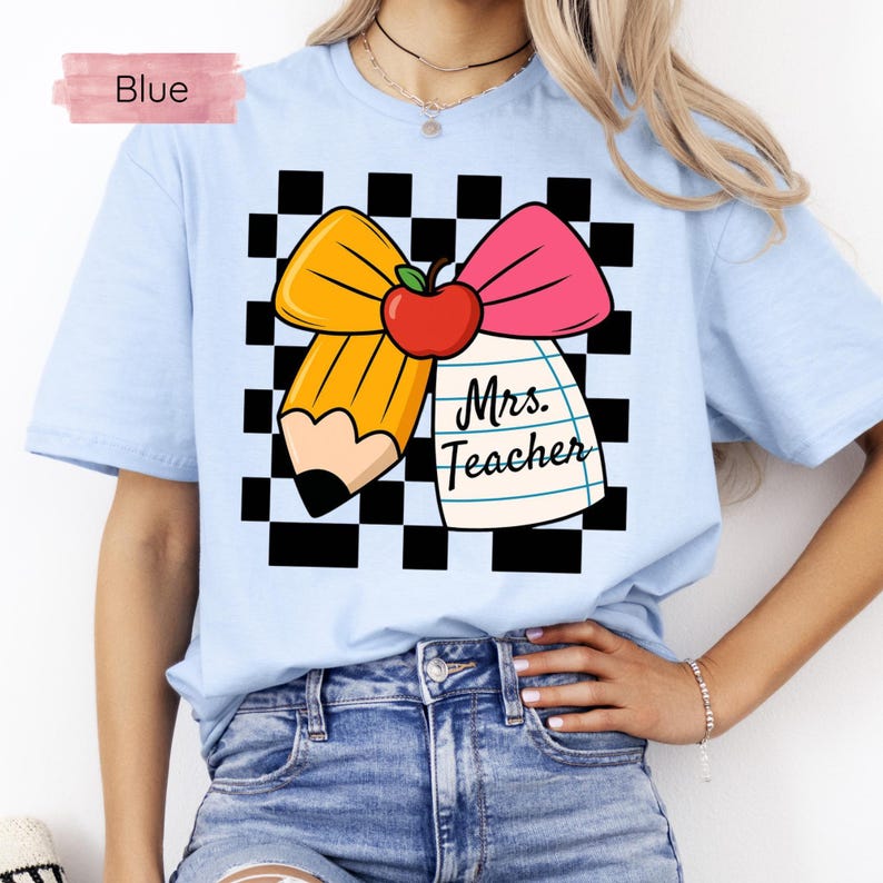 Puede incluir: Camiseta azul claro con un gr&aacute;fico que presenta un l&aacute;piz amarillo, un lazo rosa y una manzana roja sobre un fondo a cuadros blanco y negro. El texto "Mrs. Teacher" est&aacute; impreso en un bloc de notas. La palabra "Blue" est&aacute; en un rect&aacute;ngulo rosa.
