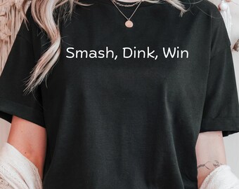 May include: Black t-shirt with the text "Smash, Dink, Win" in white. The shirt is a crew neck style and appears to be made of a soft material. The text is centered on the front of the shirt.