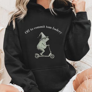 May include: Black hoodie with a humorous design. It shows a frog in a wizard's hat on a scooter, with the text "Off to commit tom foolery" above. The hoodie has a front pocket and a drawstring hood, perfect for casual wear.