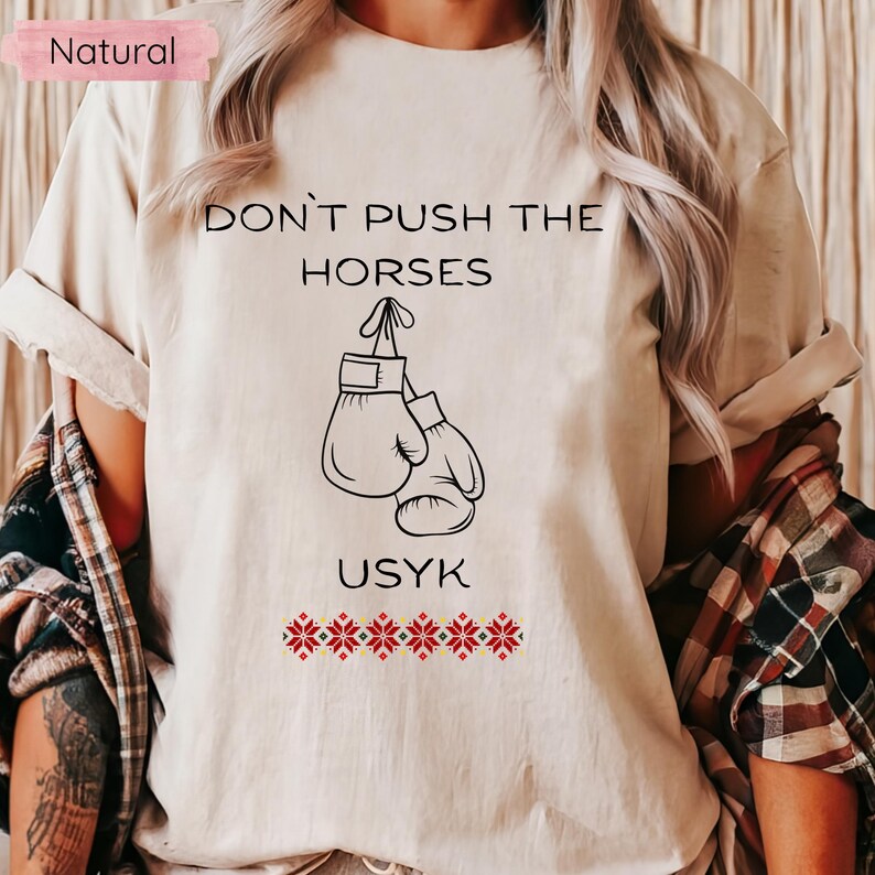 May include: A natural-colored t-shirt featuring the text "DON'T PUSH THE HORSES" above a graphic of boxing gloves. Below the gloves, the name "USYK" is printed, with a row of red and white floral designs beneath it.