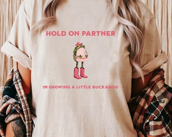 Może przedstawiać: Beżowa koszulka z różowym napisem "HOLD ON PARTNER" i "IM GROWING A LITTLE BUCKABOO". Koszulka przedstawia kreskówkową żabę w kapeluszu kowbojskim i różowych butach.