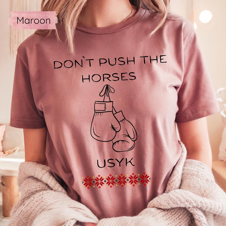 May include: Maroon t-shirt with the text "DON'T PUSH THE HORSES" above a graphic of boxing gloves. The word "USYK" is below the gloves, and a decorative pattern is at the bottom. The shirt is a solid maroon color.