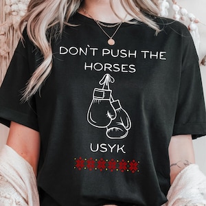 May include: Black t-shirt with the text "DON'T PUSH THE HORSES" above a graphic of boxing gloves. The name "USYK" is printed below the gloves, with a red and white patterned design at the bottom. The shirt has a crew neck.